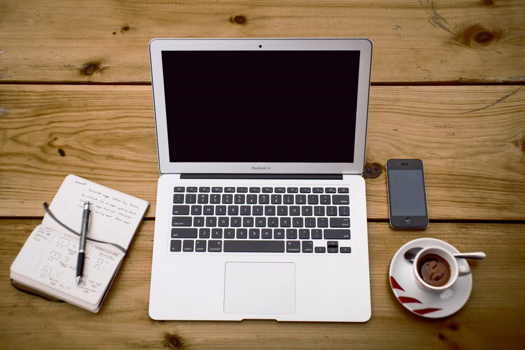 A laptop surrounded by a notebook, phone and coffee